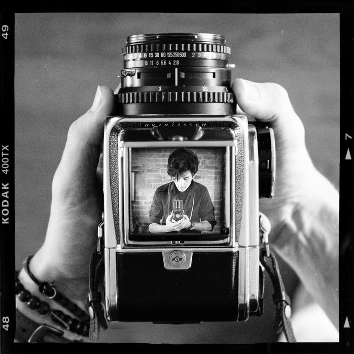 A person holds a camera with a reflection of a man holding a camera visible in its viewfinder.