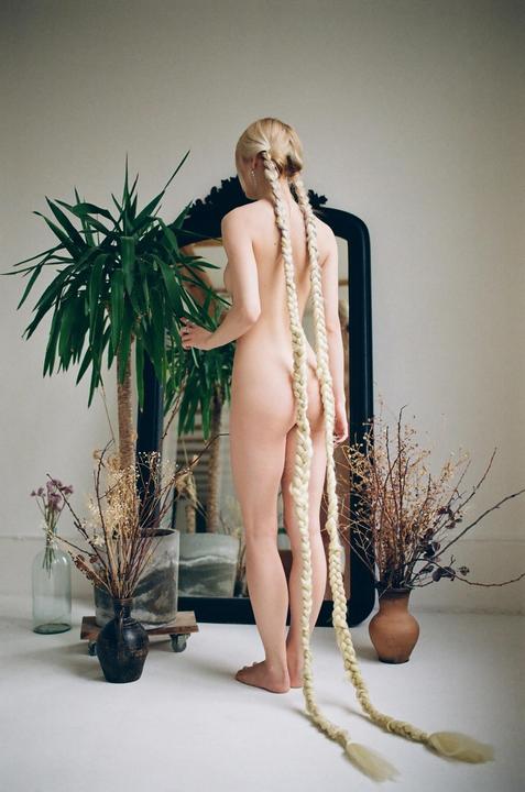 A nude woman with long braided hair stands with her back to the camera, looking into a mirror next to a large plant.