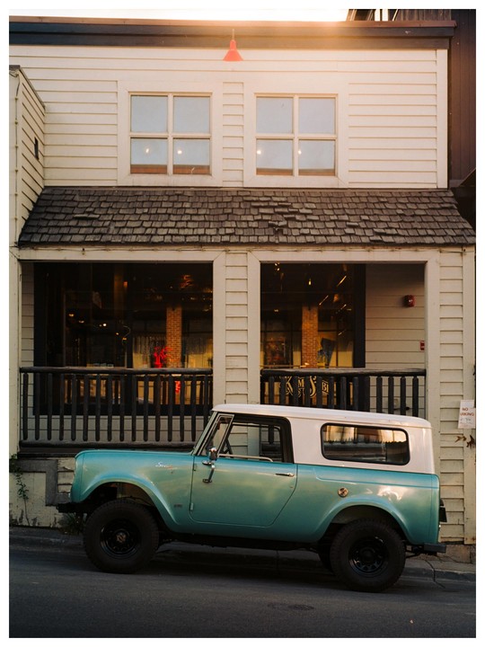 A teal and white vintage SUV is parked on a street in front of a building with a wooden facade and a small balcony.