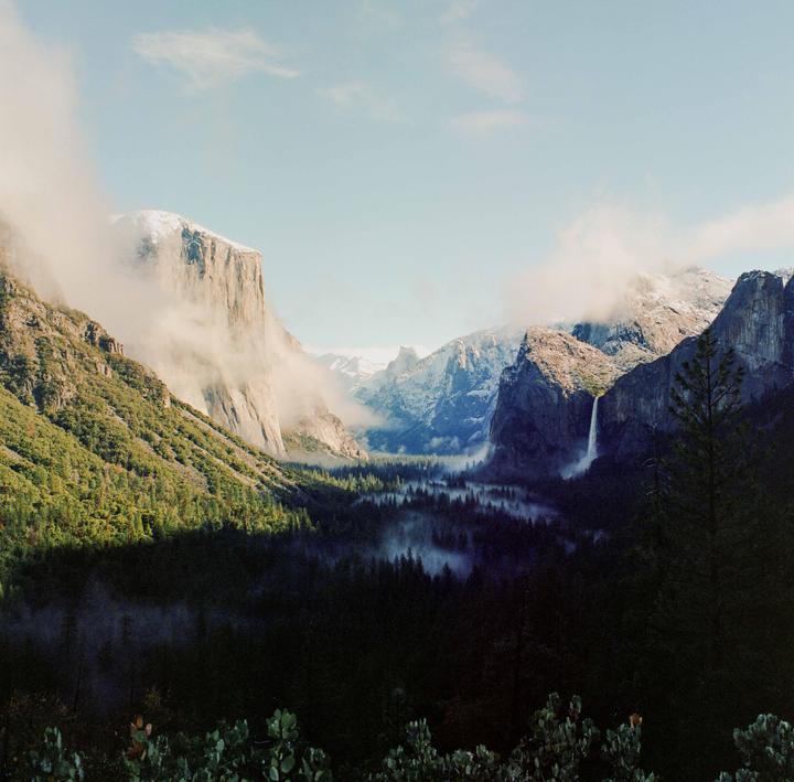 A valley with a waterfall and a forest is surrounded by mountains partially covered in fog.