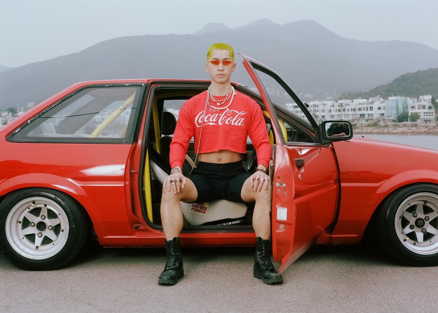 A man with yellow hair and red sunglasses sits in the open door of a red car with mountains and water in the background.