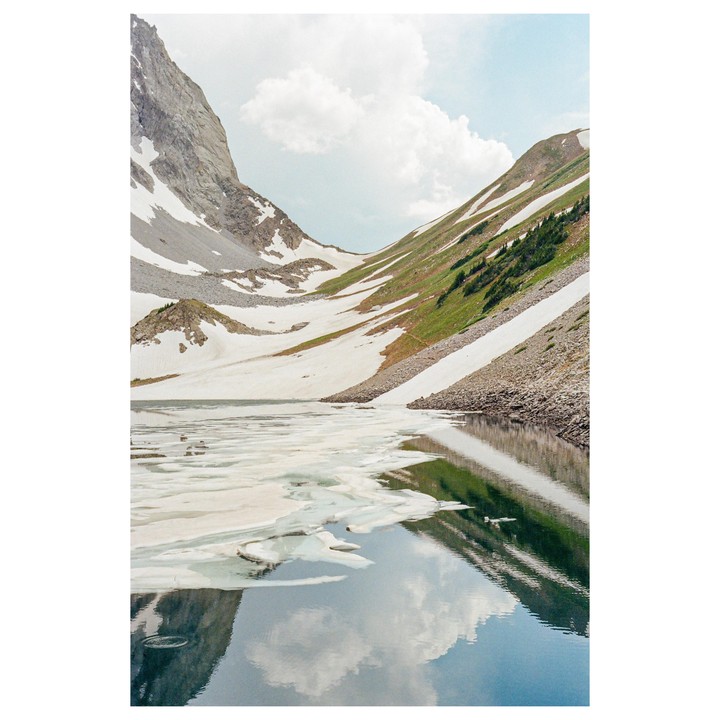 A body of water with ice floats reflects a cloudy sky and snow-covered mountains.
