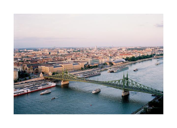 An aerial view shows a green bridge crossing a river with boats, with a city and buildings in the background.
