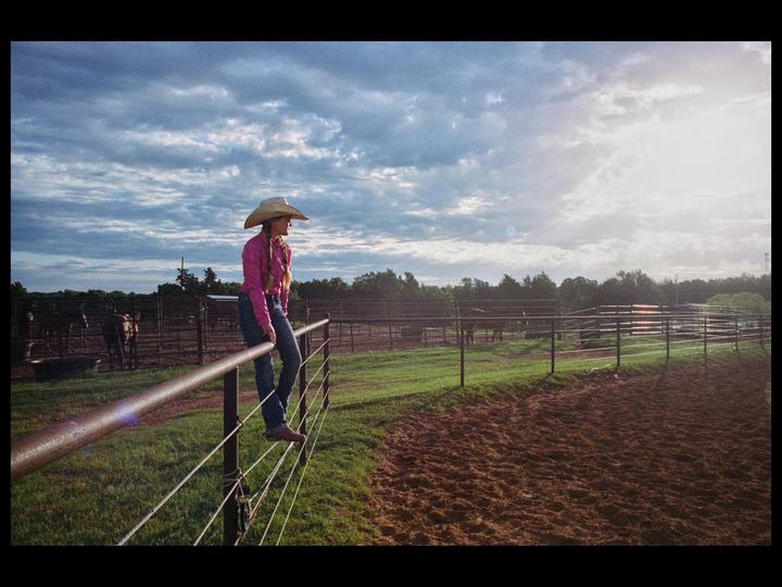 A woman in a cowboy hat and pink shirt sits on a fence at a ranch under a cloudy sky.