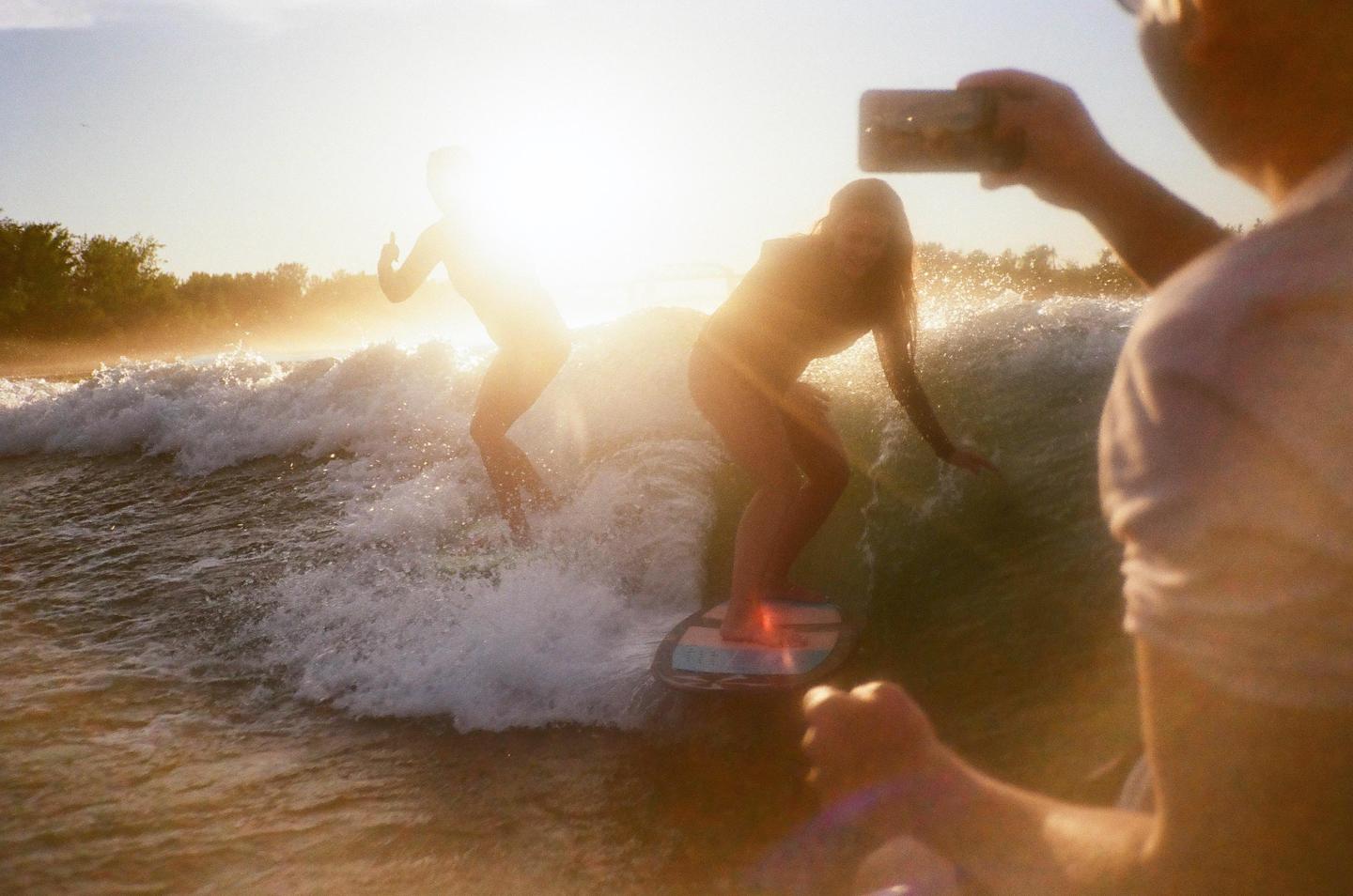 Two women are wake surfing on a boat's wake at sunset while a man films them with a phone.