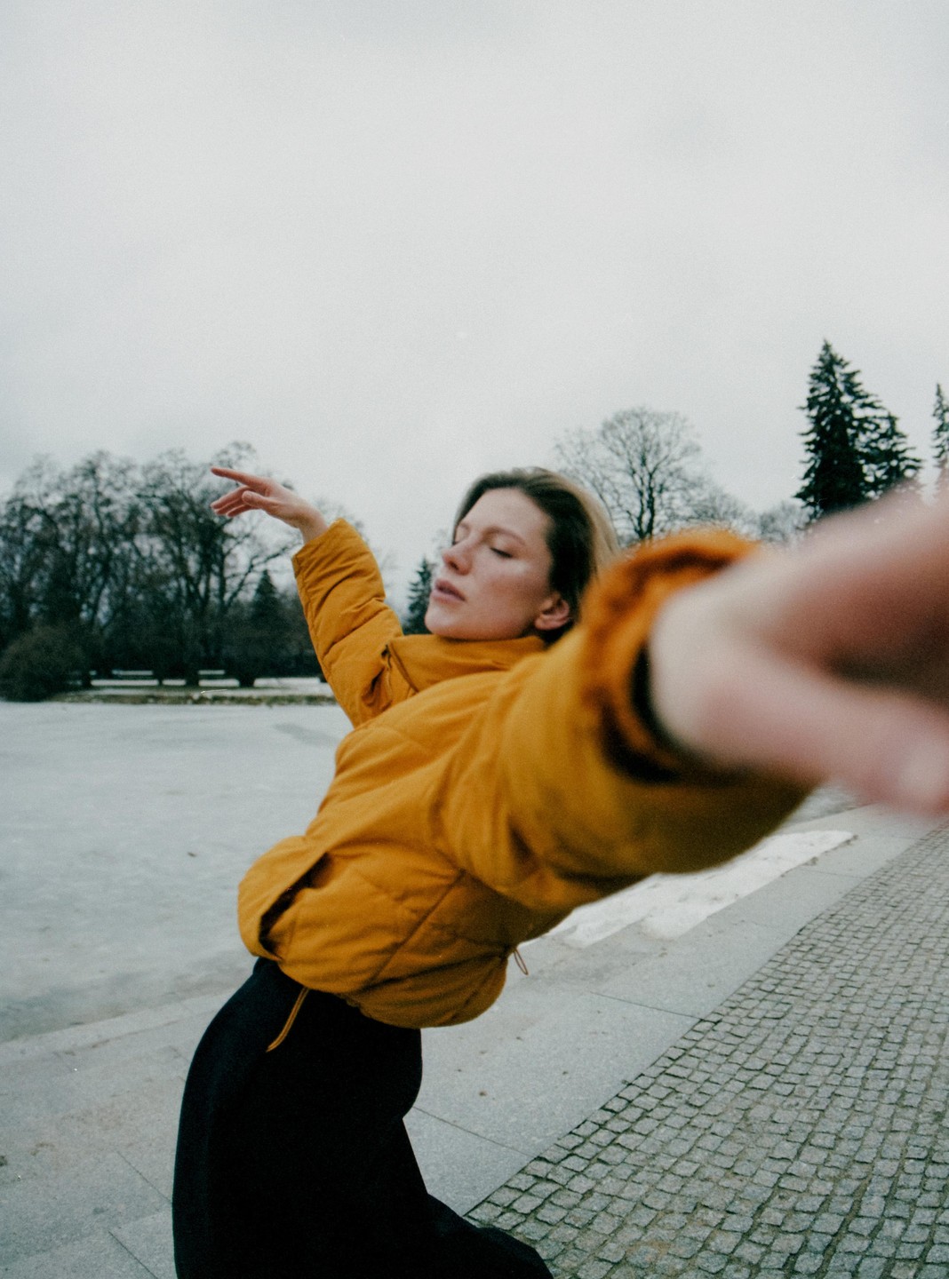 A woman in a yellow jacket extends her arm towards the camera with a frozen body of water and trees in the background.