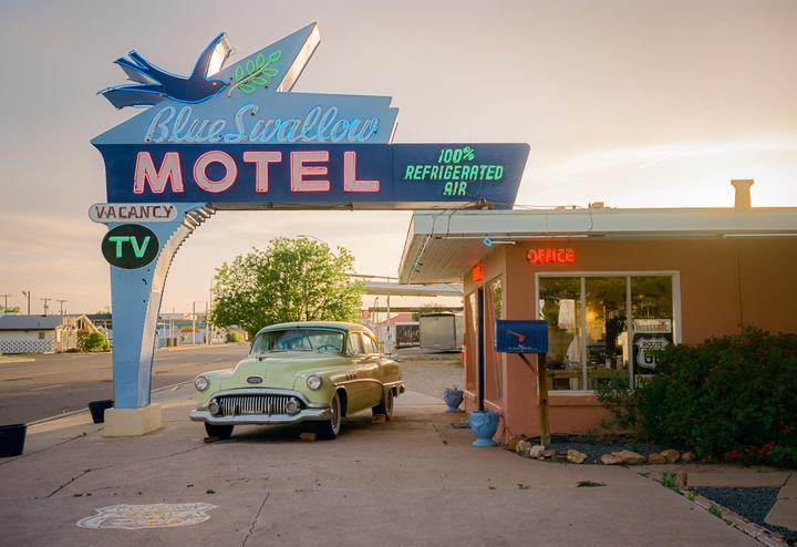 A vintage car is parked in front of the Blue Swallow Motel sign and office at sunset.