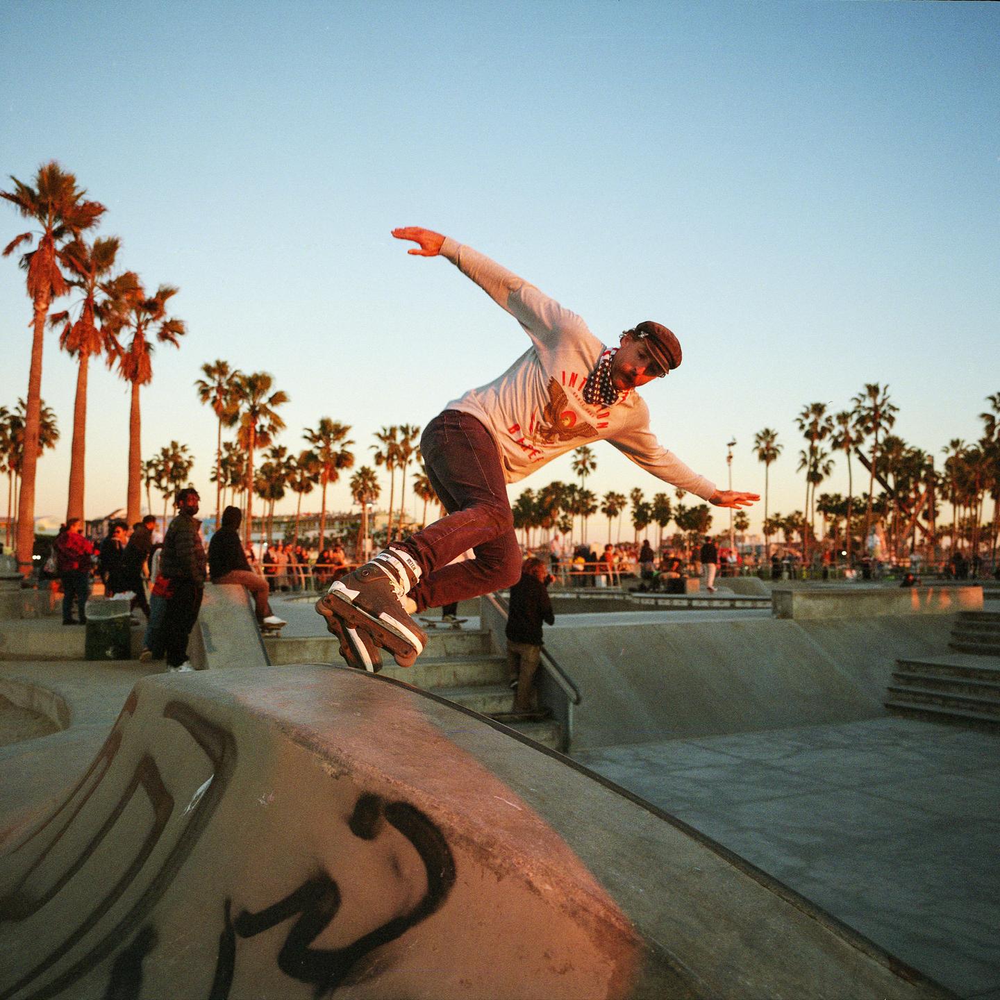 A man on rollerblades jumps over a concrete ramp in a skate park at sunset.