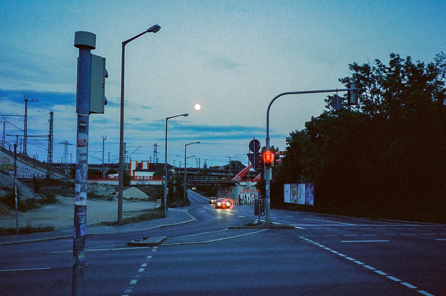 A full moon hangs in the sky over a street intersection with a red traffic light and a car approaching in the distance.