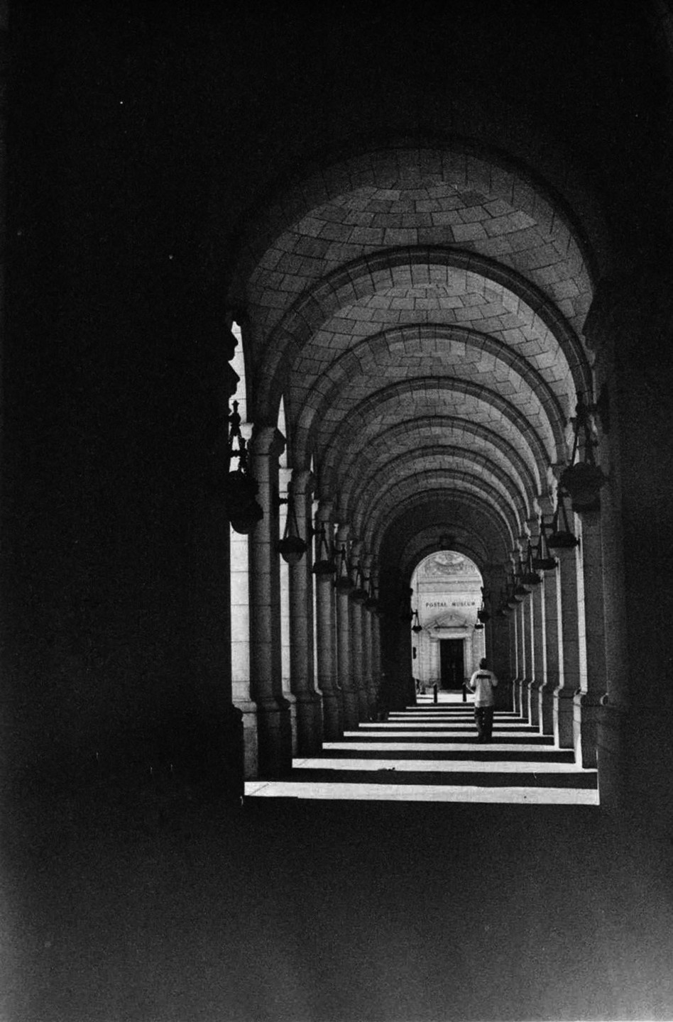 A person walks down a long arched hallway with strong shadows and light.