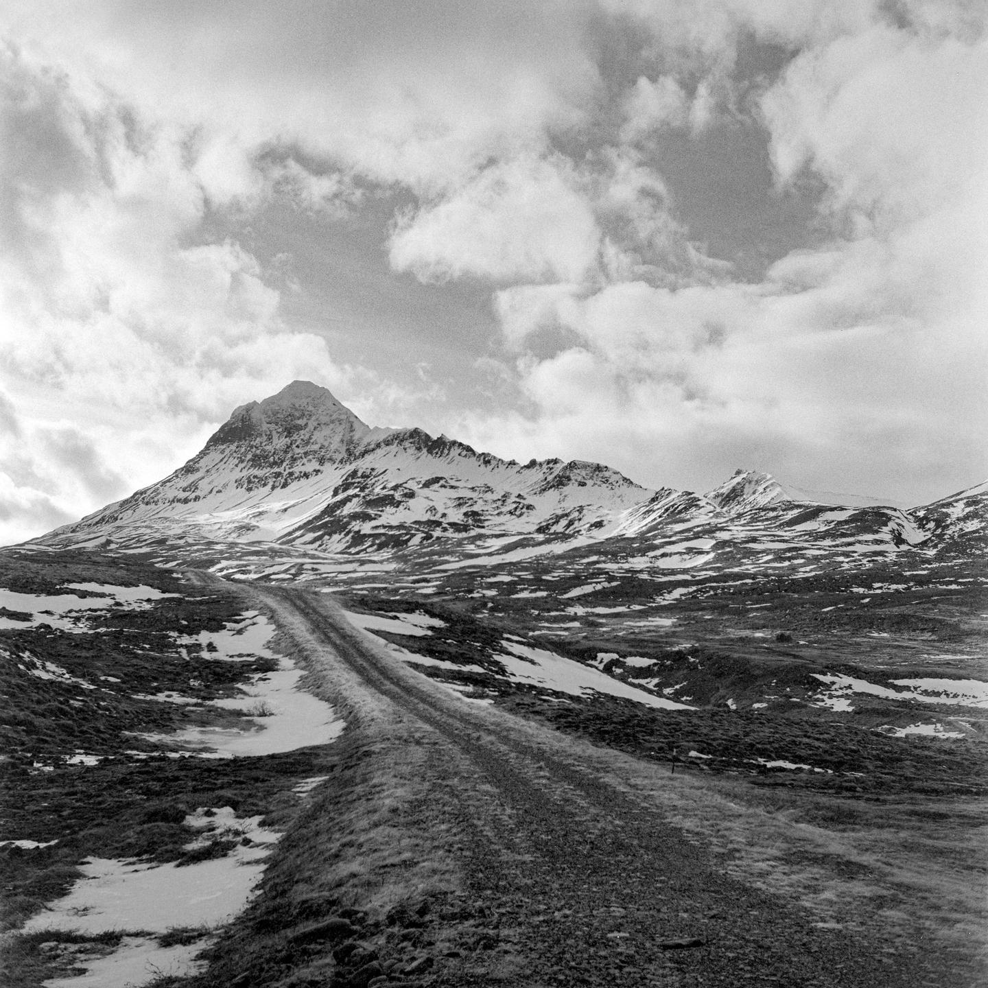 A dirt road leads towards a snow-capped mountain under a cloudy sky.