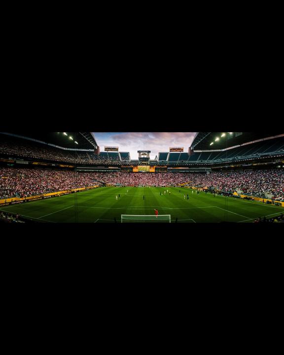 A panoramic view of a soccer match in a stadium filled with spectators under a cloudy sky.