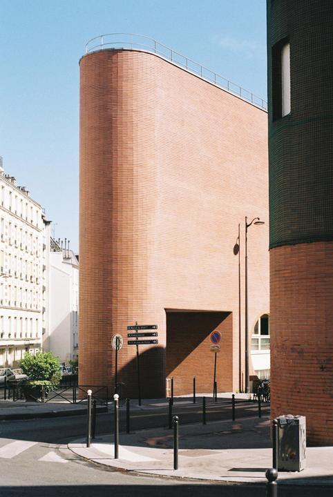 A street corner in Paris features a large, curved brick building with a smaller, darker cylindrical building next to it.