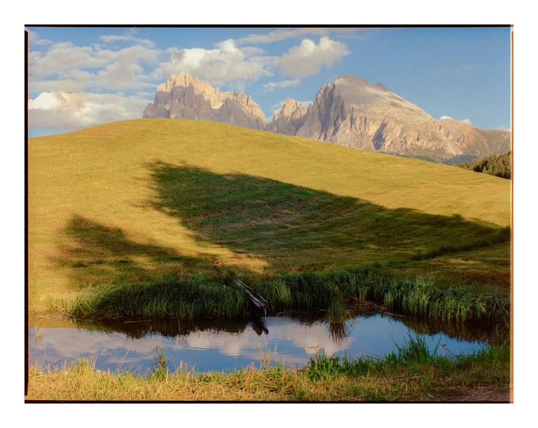 A small pond with grass around it sits in the foreground of a grassy hill with mountains in the background under a blue sky with clouds.