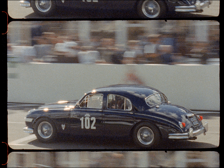 A dark blue vintage car with the number 102 on its side races past a blurred crowd.