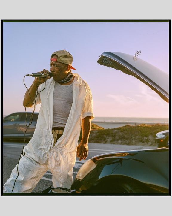 A man in a white jumpsuit sings into a microphone next to a car with its hood open, with the ocean in the background.