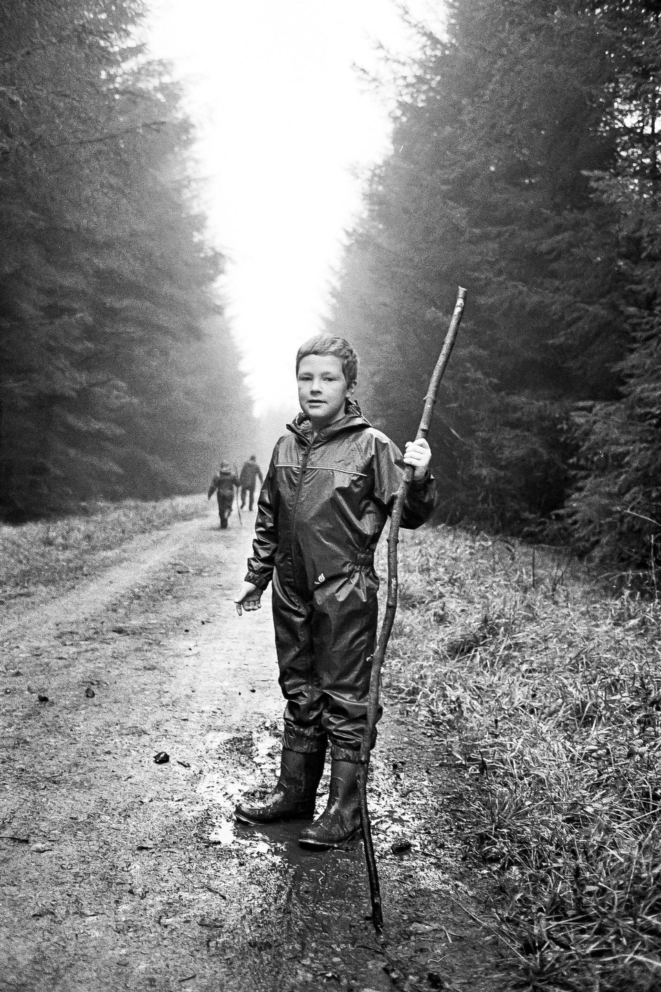 A boy in a waterproof suit and boots stands on a muddy path in a forest, holding a long stick, with two figures walking in the distance.