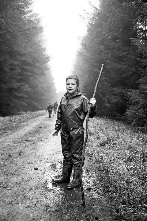A boy in a waterproof suit and boots stands on a muddy path in a forest, holding a long stick, with two figures walking in the distance.