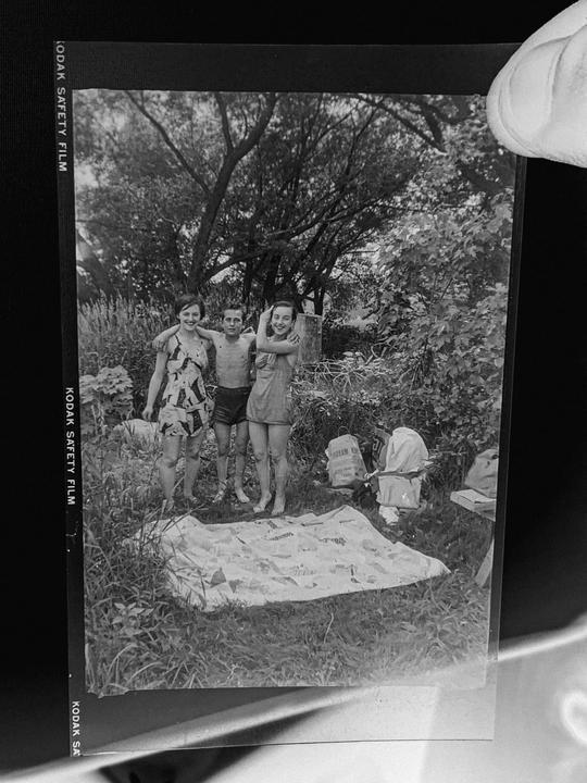 Three children stand barefoot in a grassy area with a blanket on the ground and trees in the background.