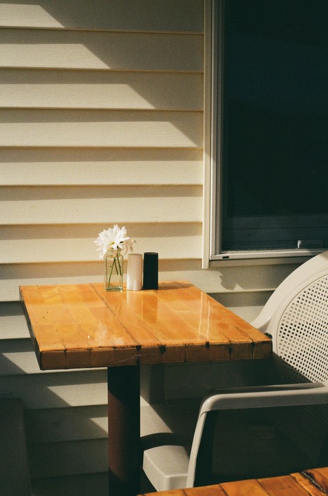 A small vase of white flowers sits on a wooden table next to a window and a chair.