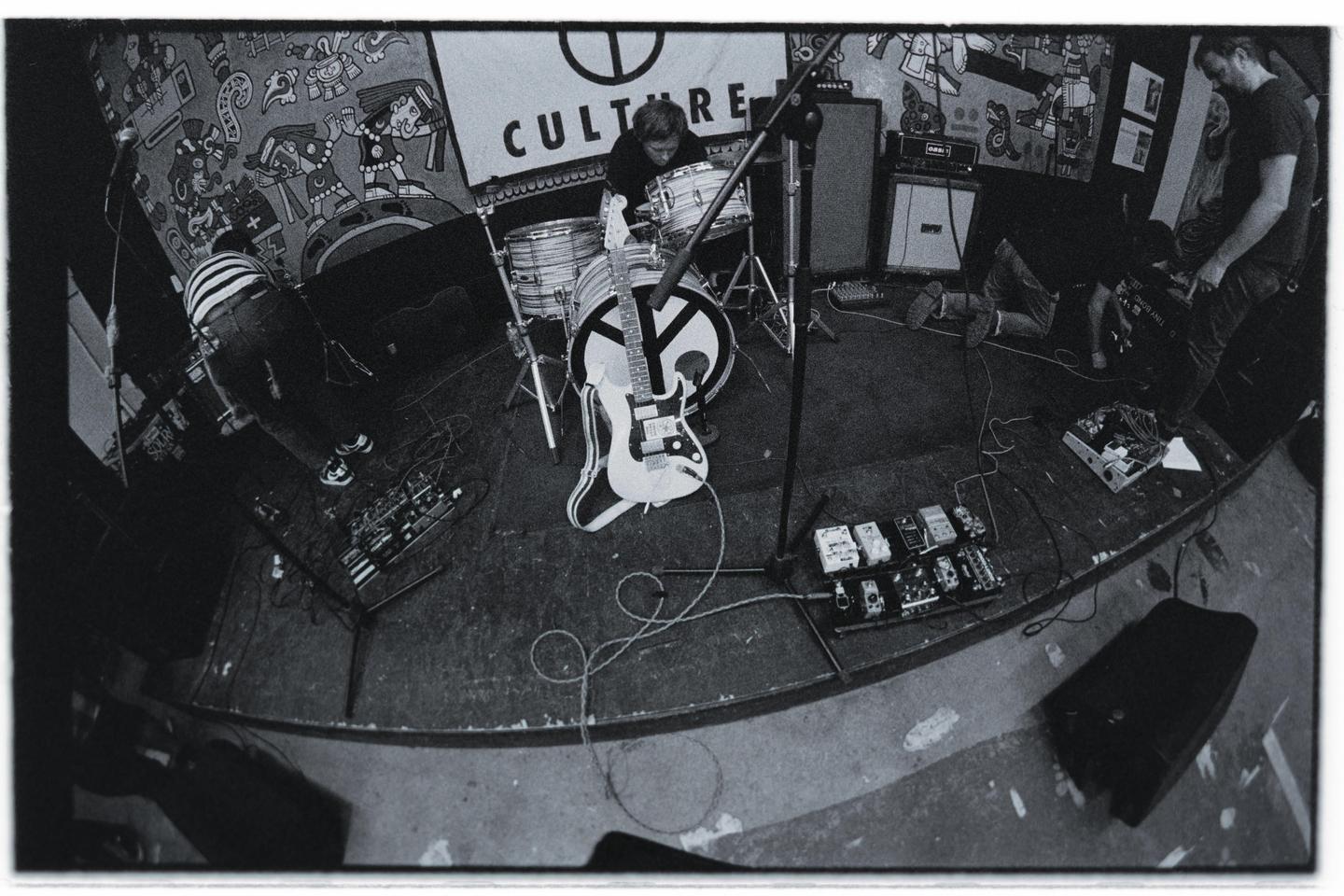 Three men are on a stage with musical instruments and equipment, including a drum kit and an electric guitar.