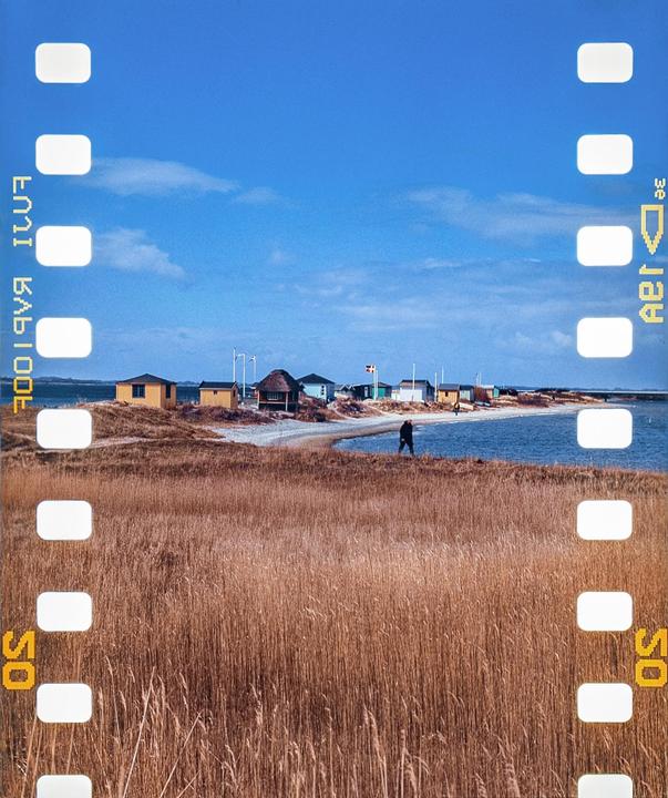 A person walks along a narrow beach with small houses and tall dry grass in the foreground.