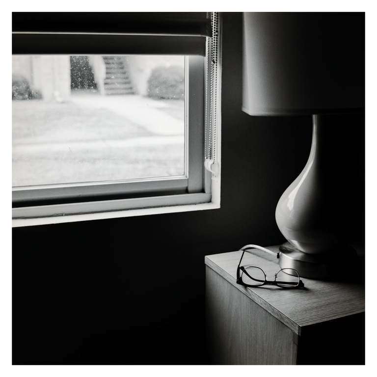 A pair of eyeglasses rests on a nightstand next to a lamp, with a window showing an outdoor view in the background.