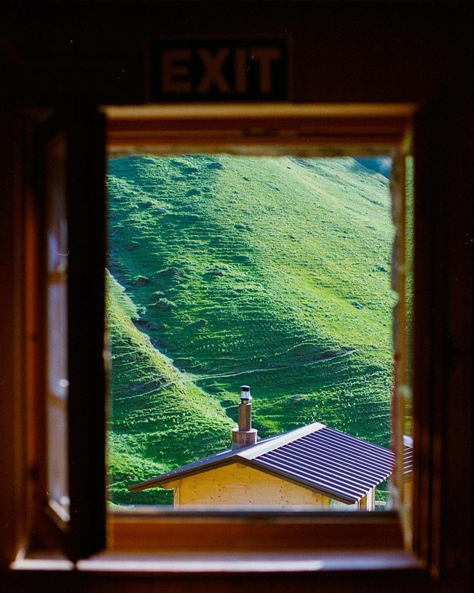 A view of a green mountain and a building roof with a chimney is seen through an open window with an "EXIT" sign above it.