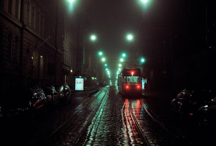 A tram with red lights approaches on a wet street at night, with green streetlights illuminating the fog.