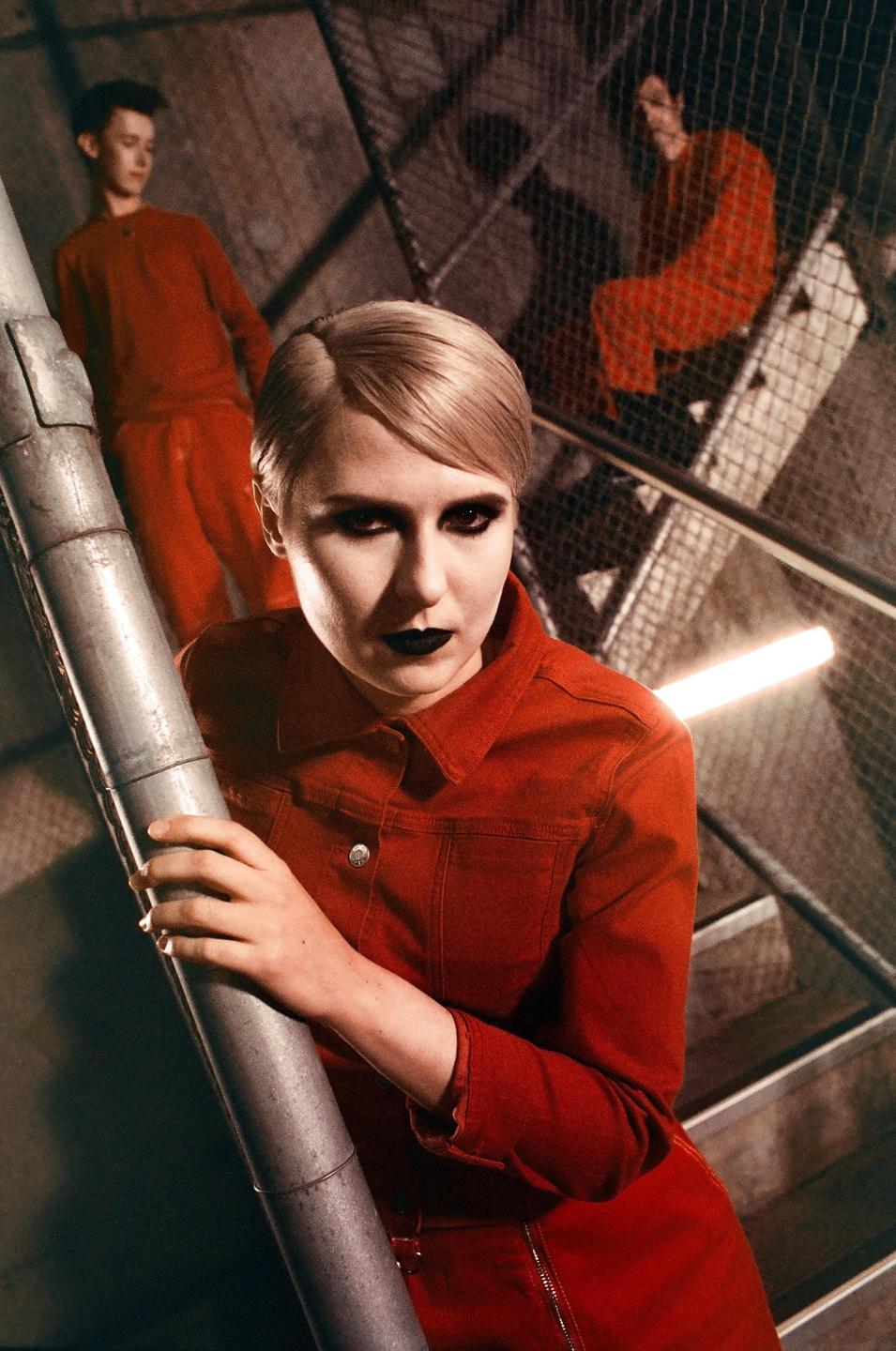 A woman in a red jumpsuit with dark makeup holds onto a metal pole while two men in red outfits stand on a metal staircase behind her.