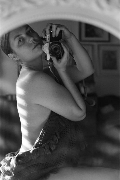 A woman photographs herself in a mirror, wrapped in a blanket with light stripes across her back.