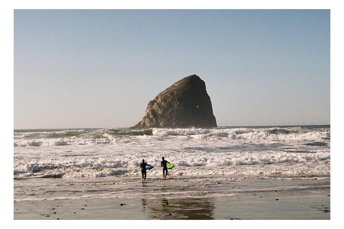 Two surfers with bodyboards walk into the ocean with a large rock formation in the background.
