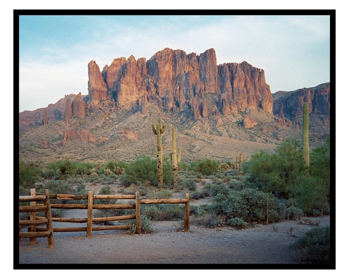 A wooden fence stands in a desert landscape with saguaro cacti and mountains under a clear sky.