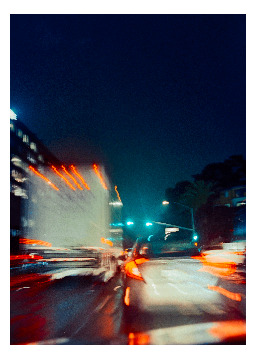 A blurry night street scene shows the red and white streaks of vehicle lights and a traffic light.