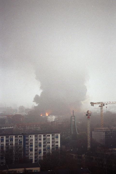 A large plume of smoke rises above a city with visible flames and construction cranes.