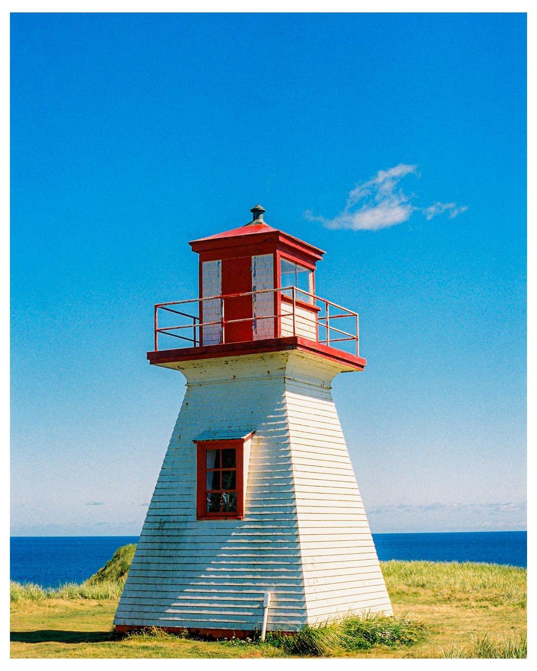 A white and red lighthouse stands on a grassy cliff overlooking the ocean under a clear blue sky.