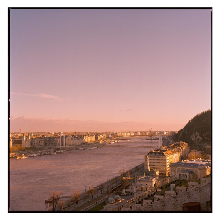 A wide view of Budapest shows the Danube river, bridges, and buildings along the riverbank, with a hill on the right.