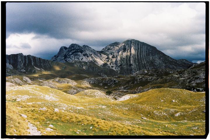 A dramatic mountain landscape features rolling hills in the foreground and rugged peaks under a cloudy sky.