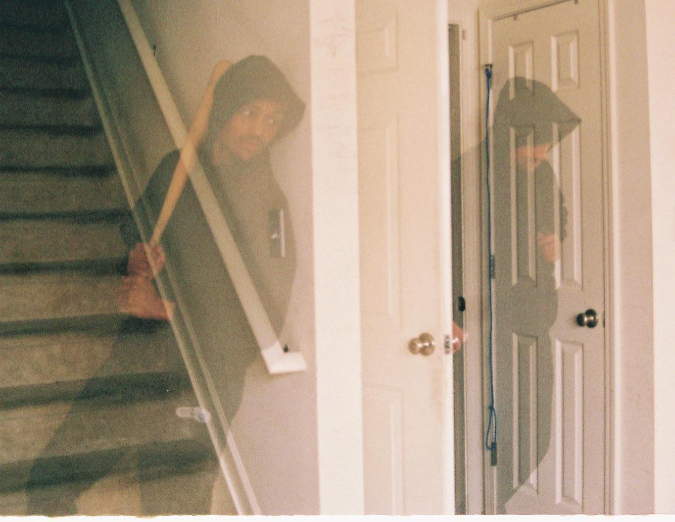 A man in a hooded sweatshirt is visible through a double exposure, once on a staircase and once as a shadow on a door.