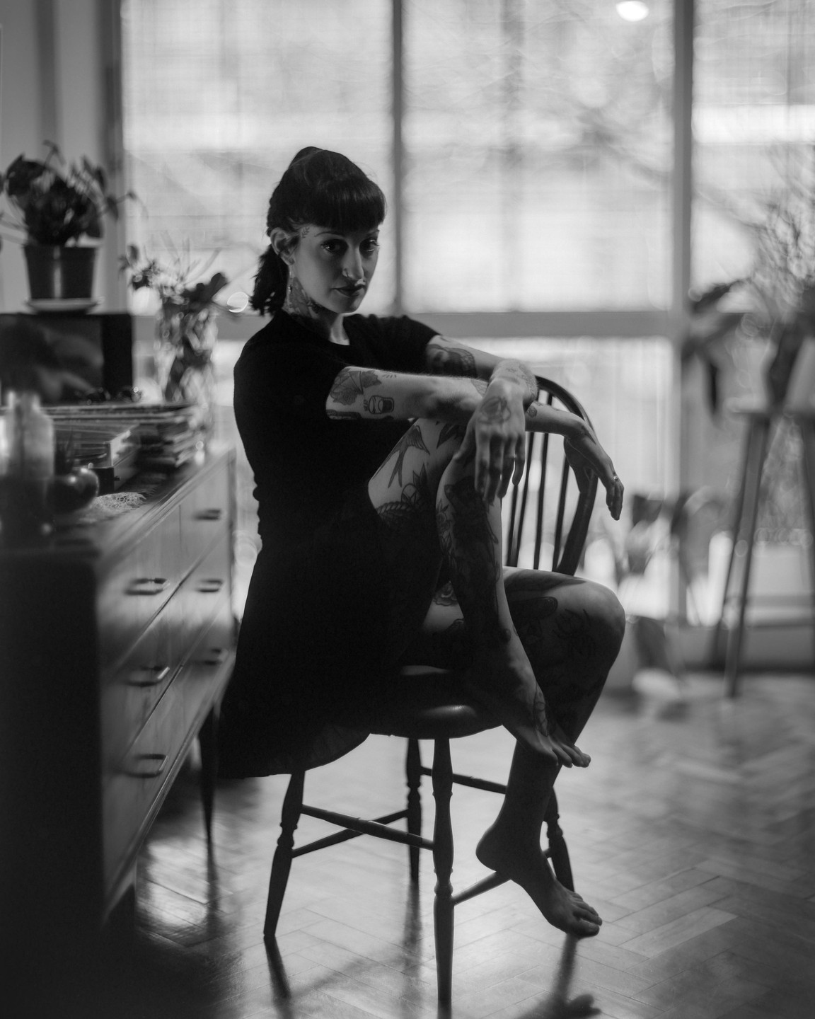 A woman with tattoos sits on a wooden chair in a room with a large window.