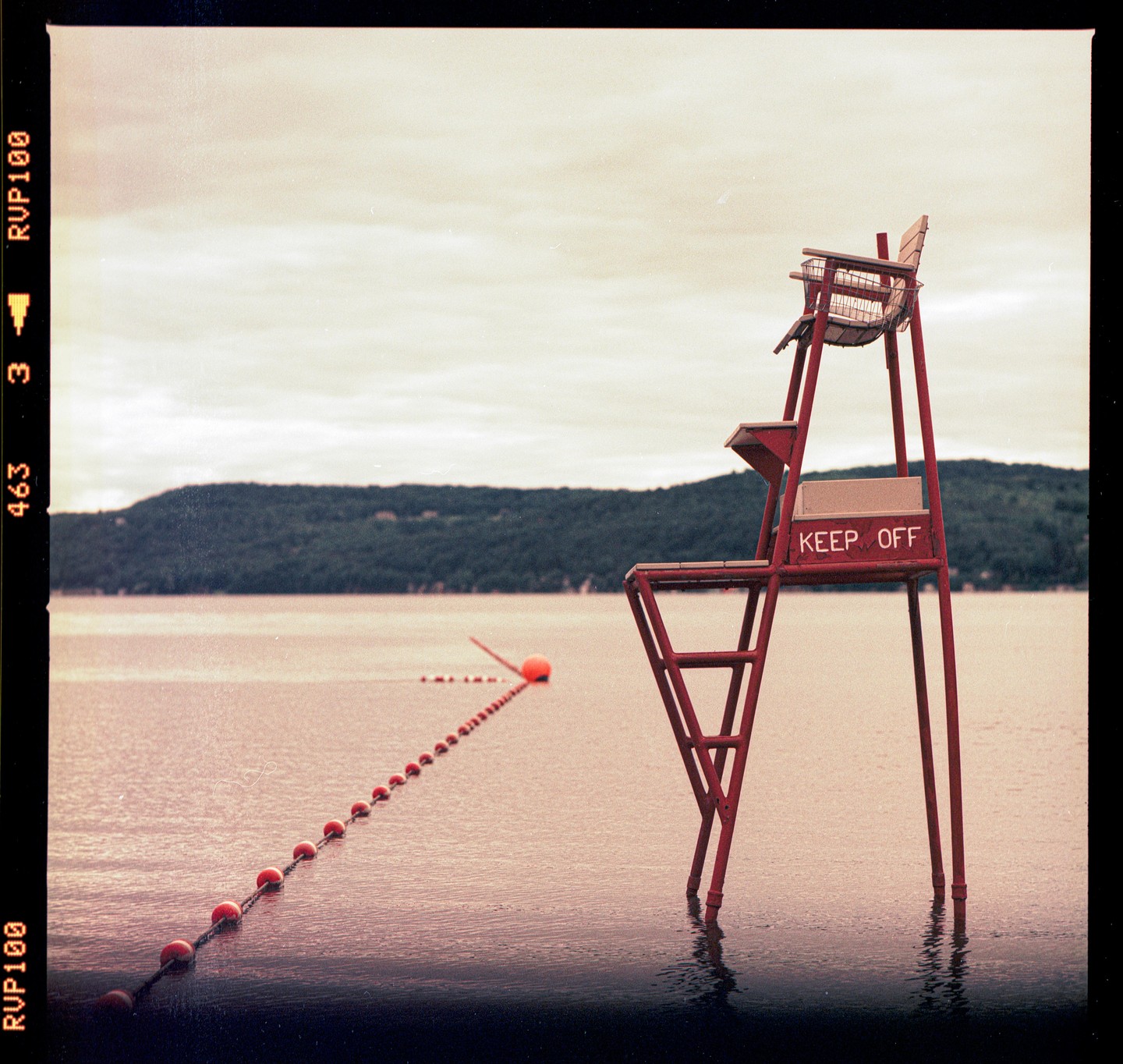 A red lifeguard chair stands in the water next to a line of buoys with a forested hill in the background.