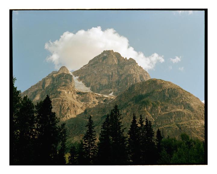 A large mountain with a cloud above its peak rises above a line of evergreen trees.