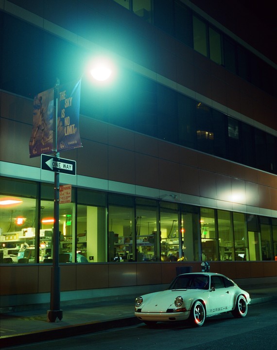 A white Porsche is parked on a street at night next to a building with many windows and a street light.