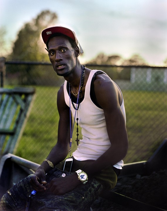 A man wearing a red baseball cap and a white tank top sits outdoors, looking directly at the viewer.