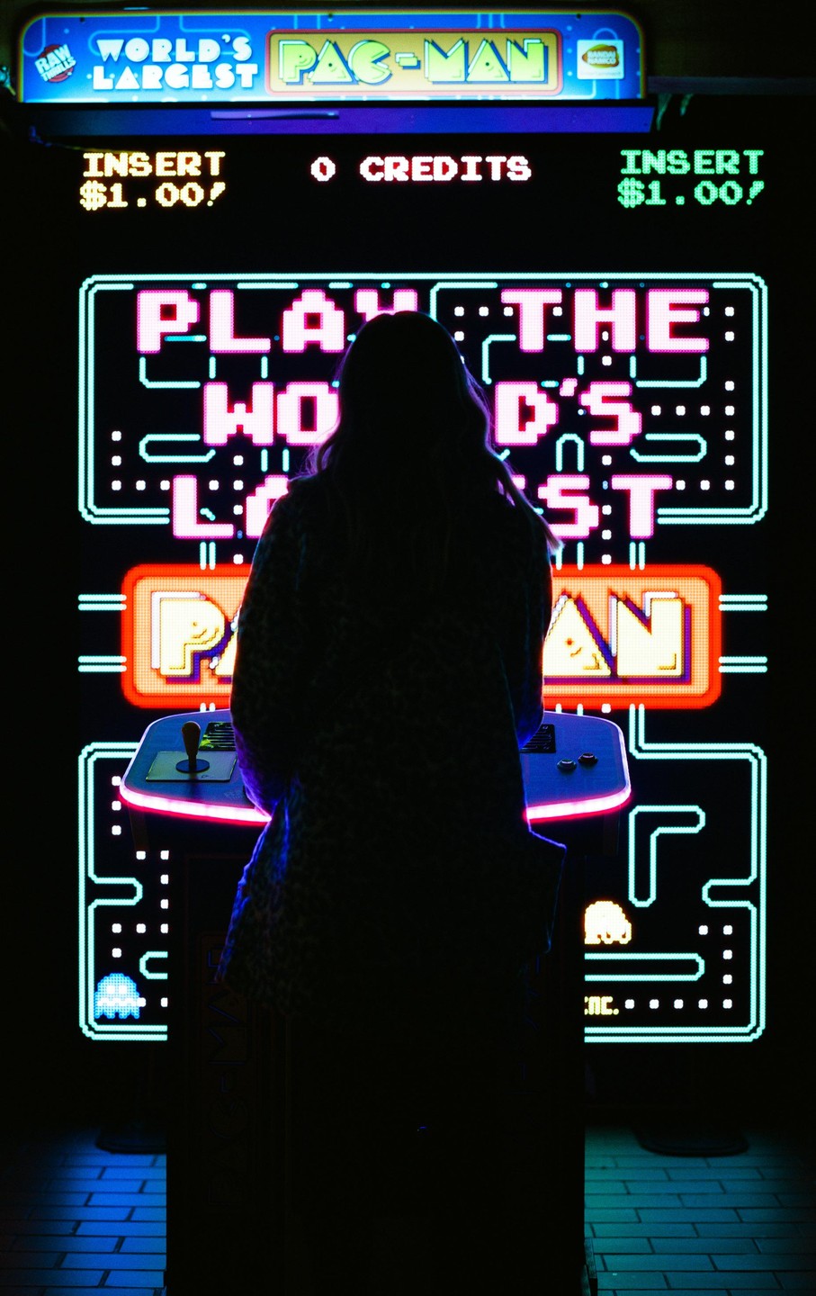 A person stands in front of a large Pac-Man arcade game, silhouetted by the neon lights.