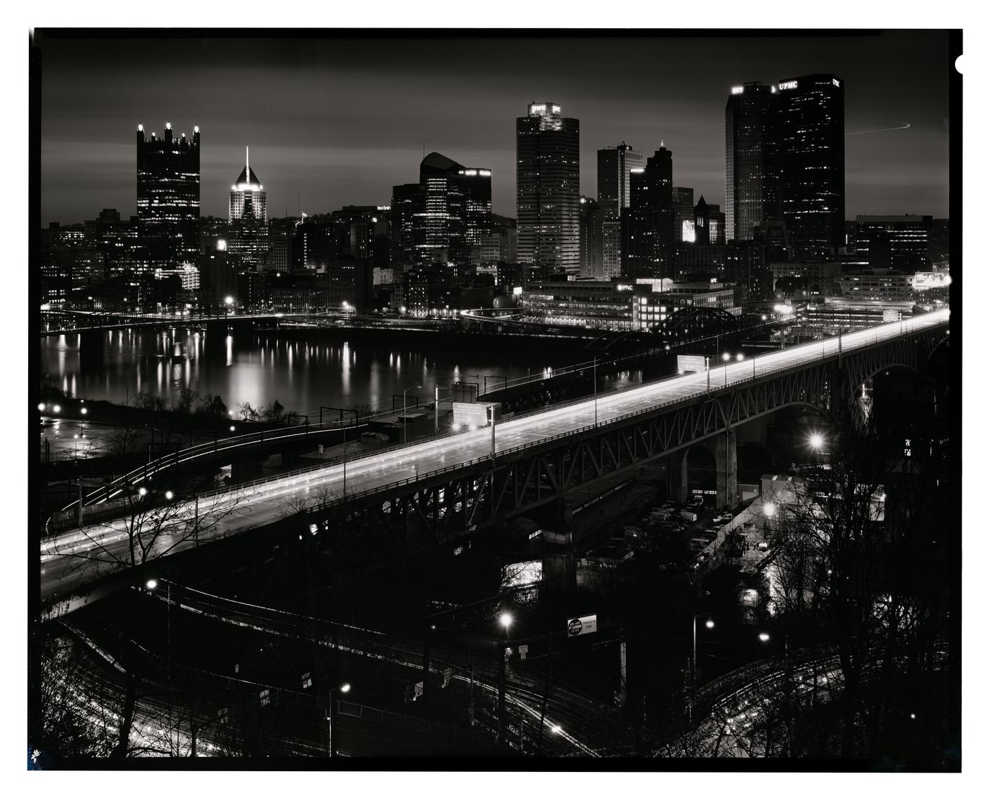 A city skyline with tall buildings and a bridge over a river is illuminated at night.