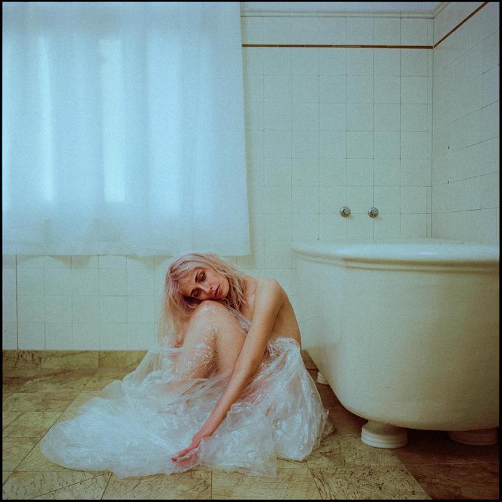 A woman with pink hair sits on the floor of a bathroom next to a bathtub, covered in plastic sheeting.
