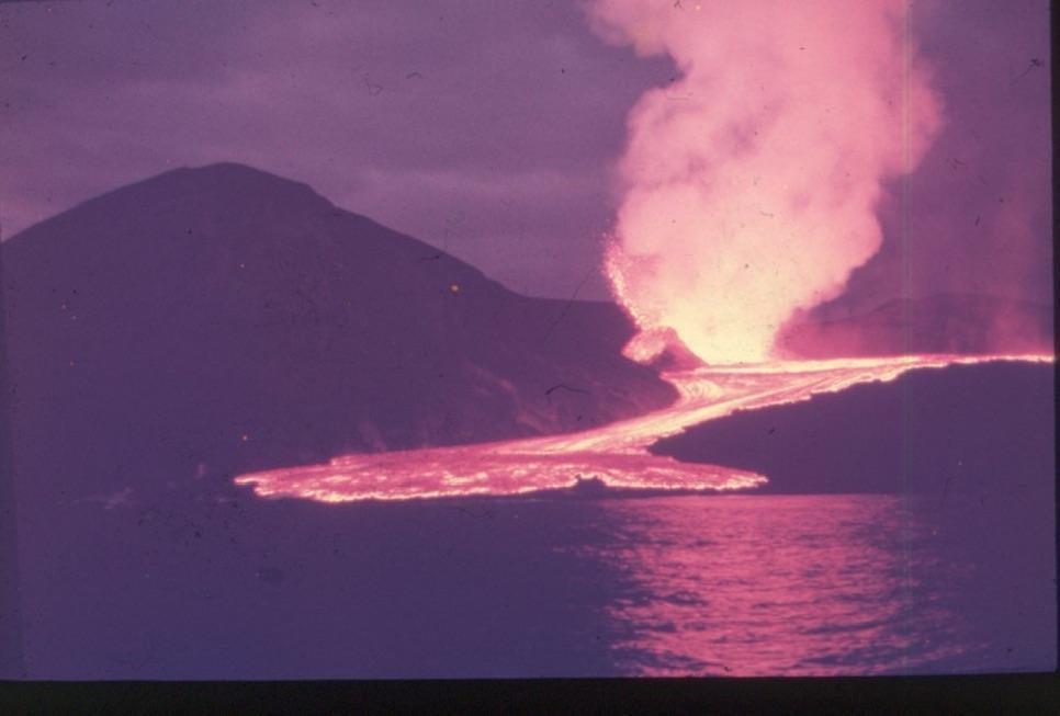 A volcano erupts, sending lava flowing down its side into the ocean at night.