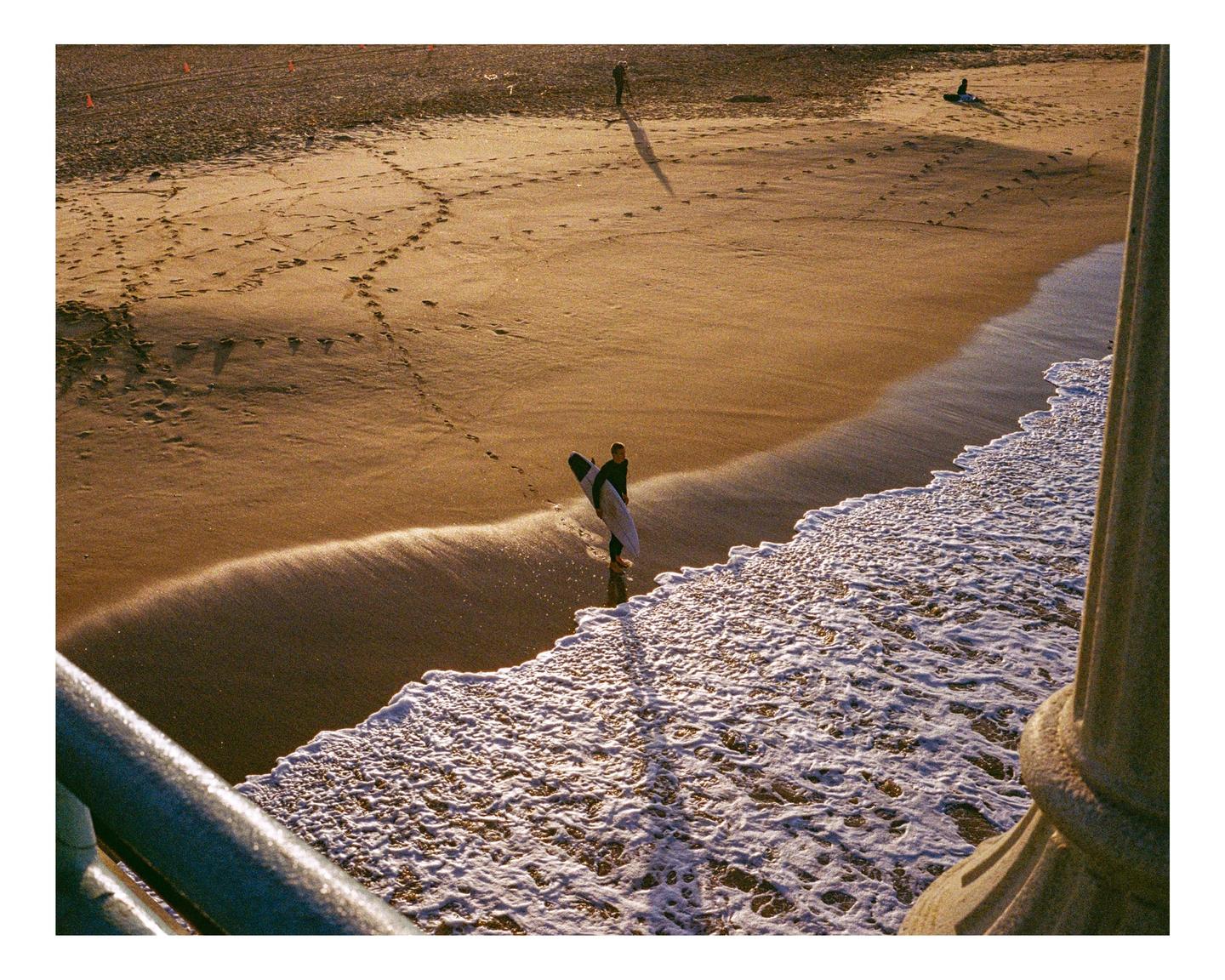 A surfer stands in the shallow water at the edge of a sandy beach, holding a surfboard.