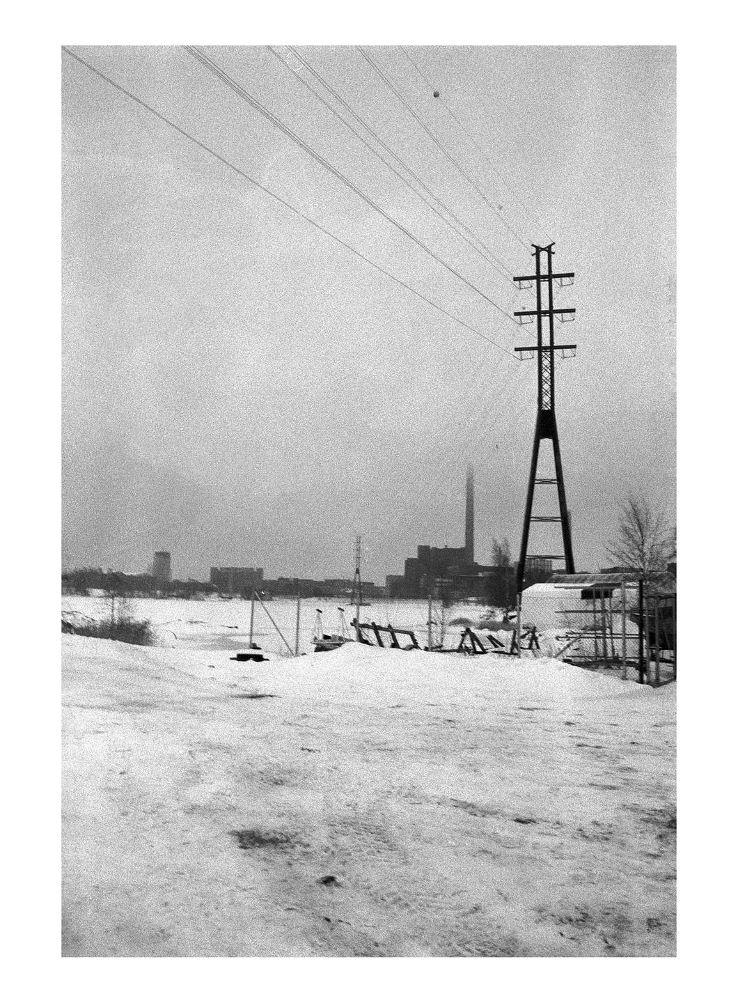 A snowy landscape features a large power line tower in the foreground with industrial buildings and a city skyline in the distance.